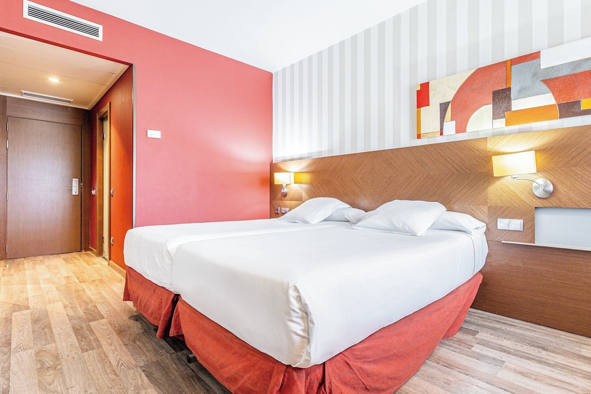 Habitación de hotel con cama doble, suelo de madera, pared de acento roja y una obra de arte abstracta moderna sobre la cama.
