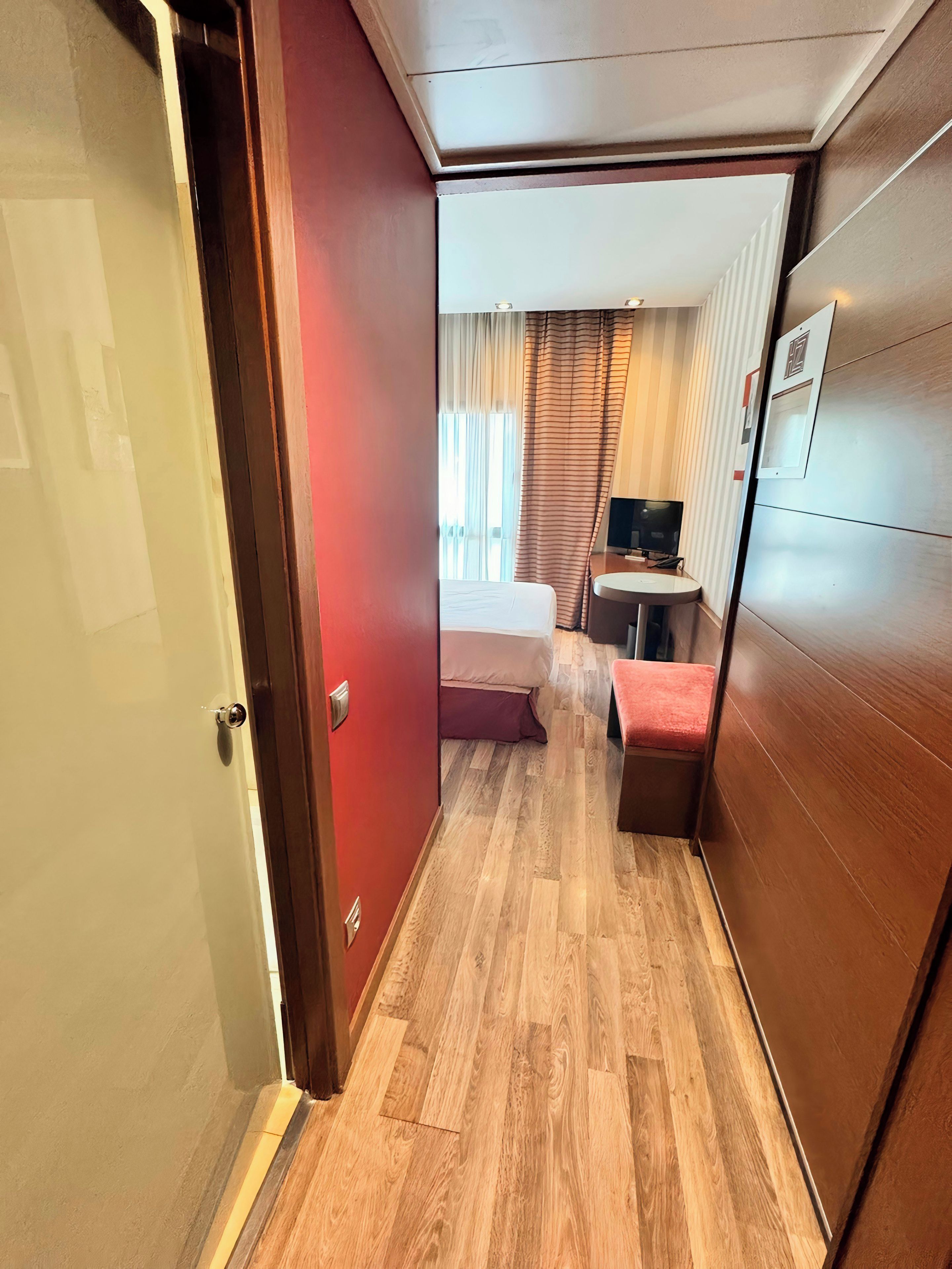 Une chambre d’hôtel avec un plancher en bois, un lit avec une tête de lit rouge, un petit bureau avec une chaise et un téléviseur.