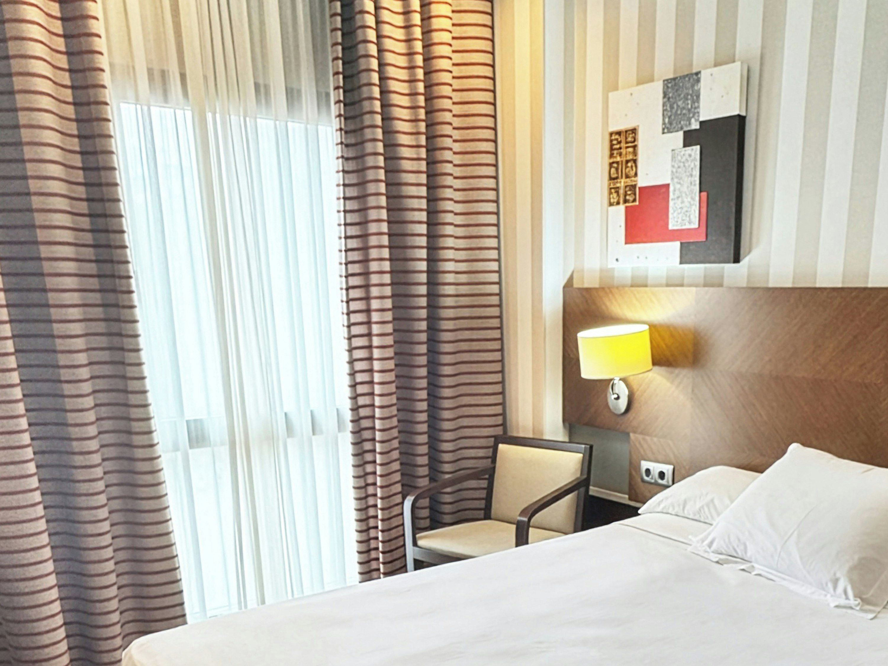 Una habitación de hotel con cama, una silla, una lámpara y cortinas.