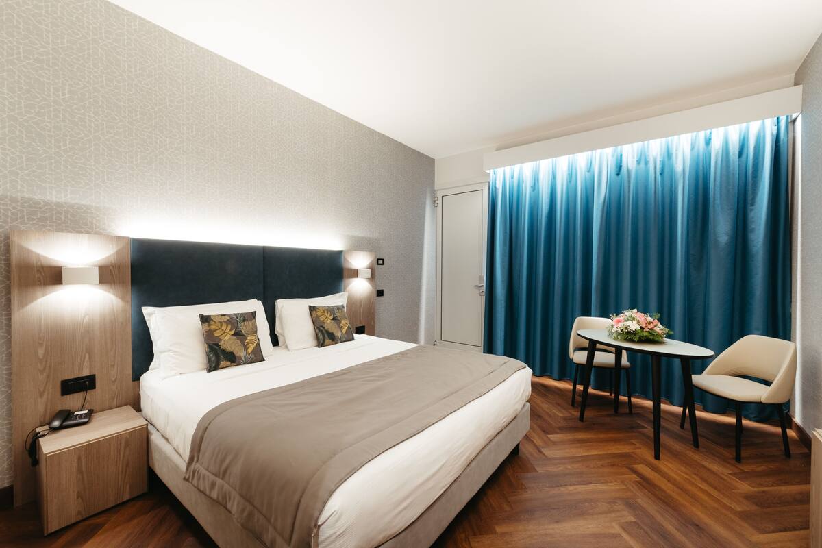 A modern hotel room with a large bed, bedside lamps, a small table with chairs, and a view of the outside through a window with blue curtains.