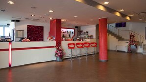 A modern hotel lobby with a reception desk, red chairs, a staircase, and a bar area.