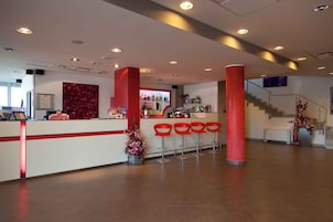 Um hall de hotel moderno com uma mesa de recepção, cadeiras vermelhas, uma escadaria e um espaço de bar.