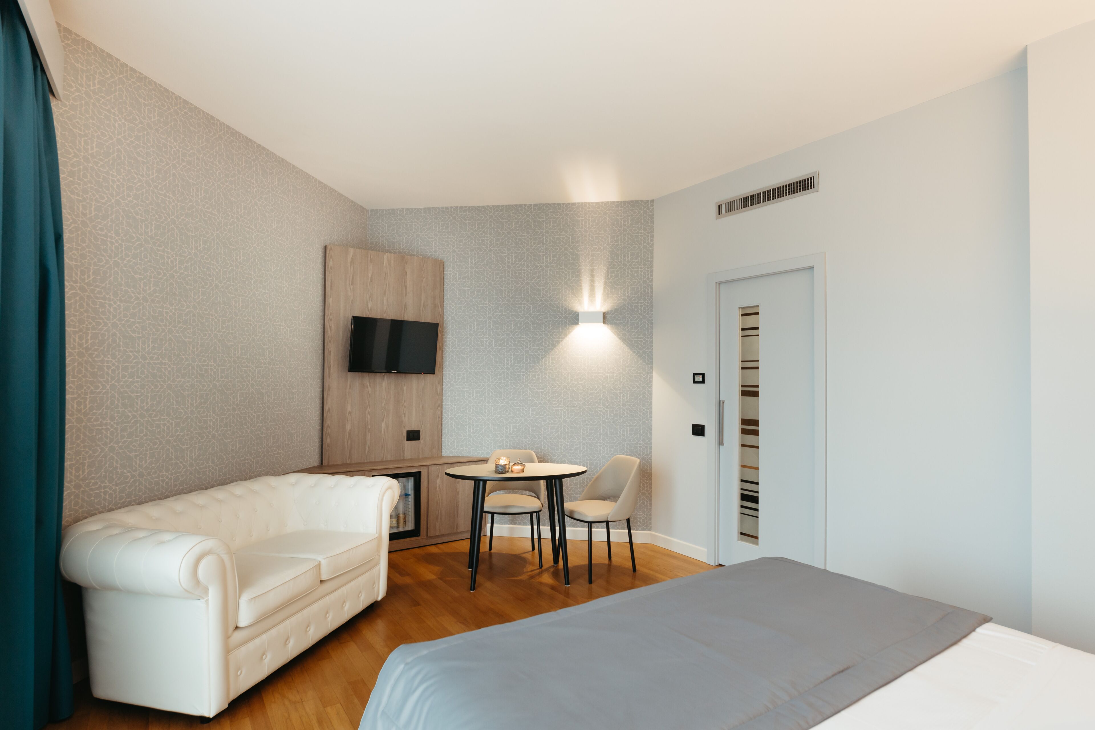 Una habitación de hotel moderna con una cama, un sofá, una pequeña mesa con sillas, un televisor montado en la pared y una puerta con ventilación.