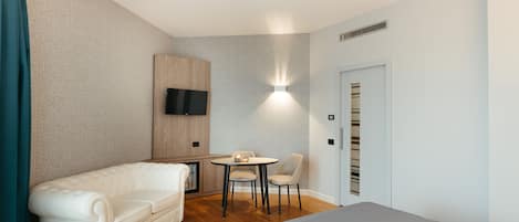A modern hotel room with a bed, a sofa, a small table with chairs, a TV mounted on the wall, and a door with a vent.