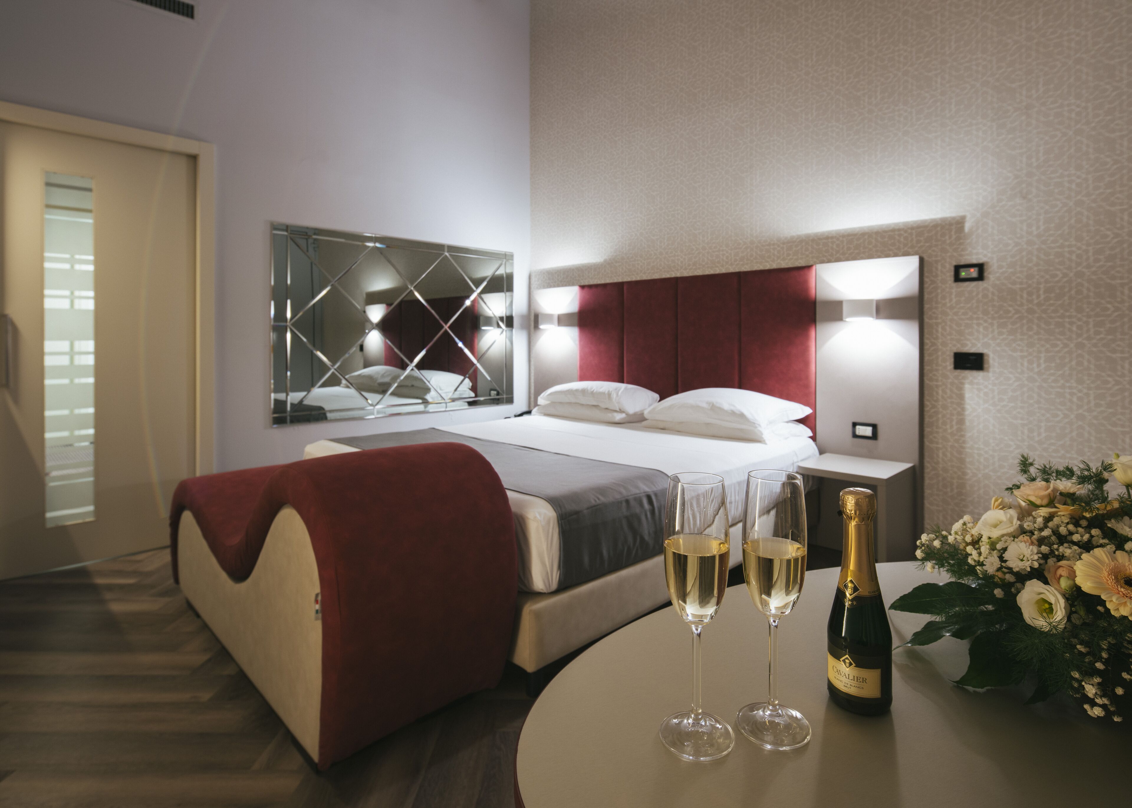A hotel room with a bed, bedside tables, a mirror, and a table with two glasses and a bottle of champagne.