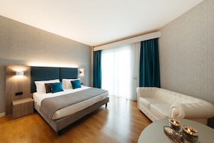 Quarto de hotel moderno com uma cama grande, um sofá branco e uma mesa de centro redonda.