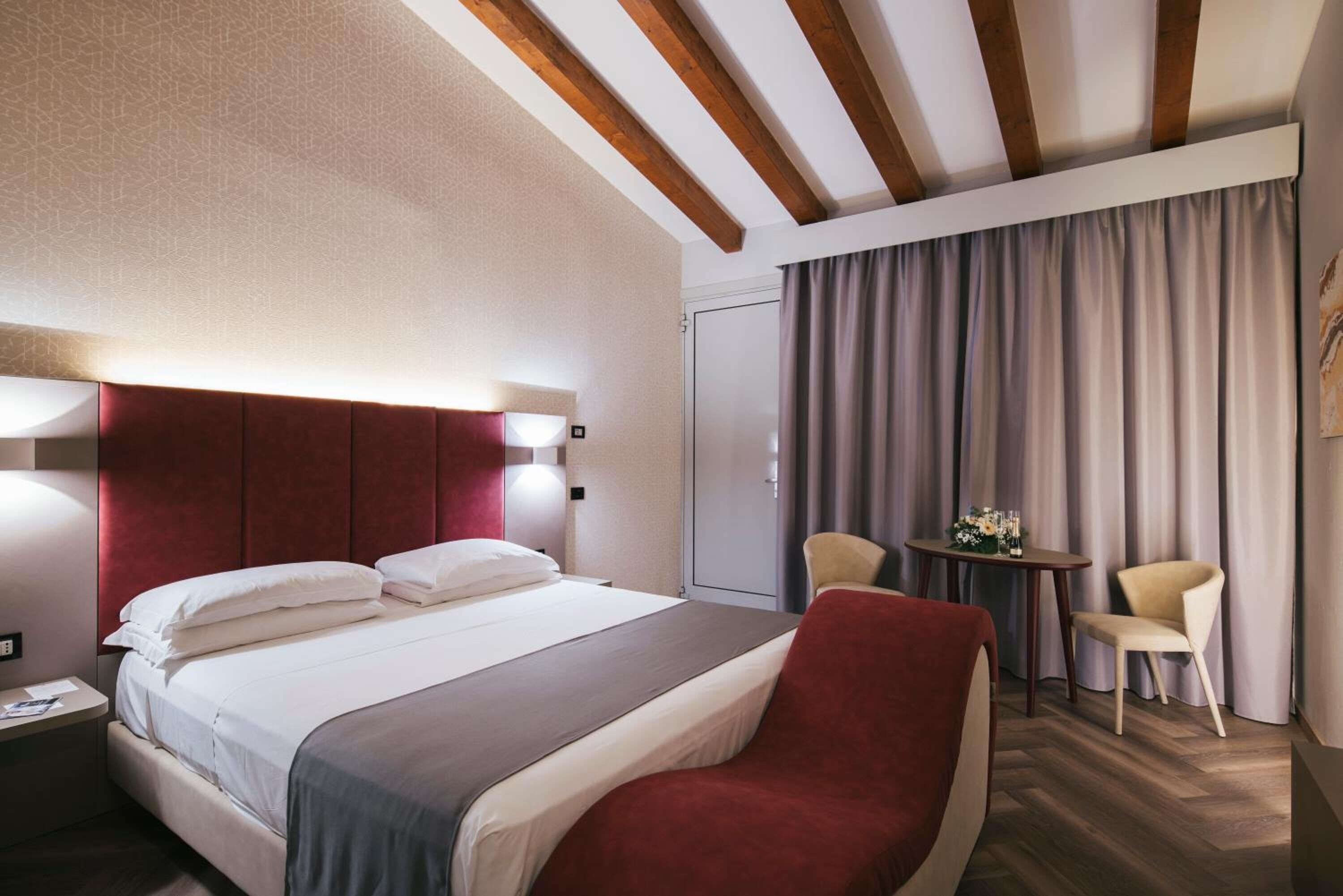 Una habitación de hotel con una cama, cabecera roja, dos lámparas de noche, una mesita con un florero, dos sillas y techo de madera.