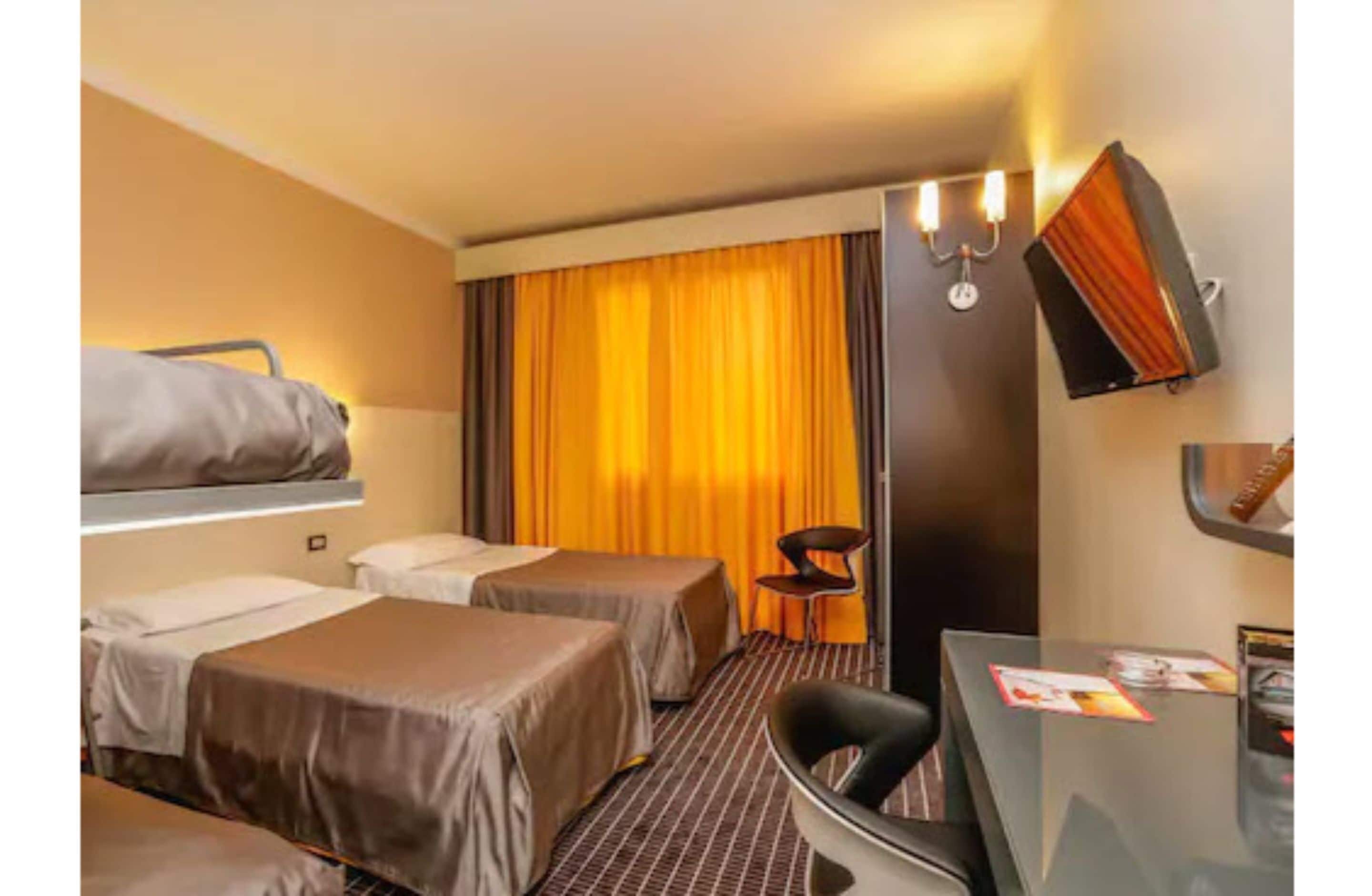 Habitación de hotel con dos camas individuales, un escritorio, una silla, un televisor y una cortina.
