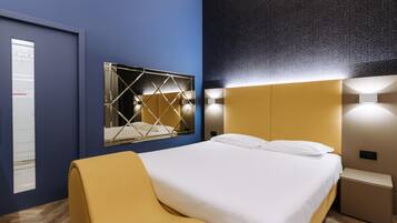 A modern hotel room with a large bed, a mirror with a geometric frame, and a wall-mounted light fixture.