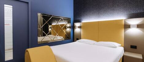 A modern hotel room with a large bed, a mirror with a geometric frame, and a wall-mounted light fixture.
