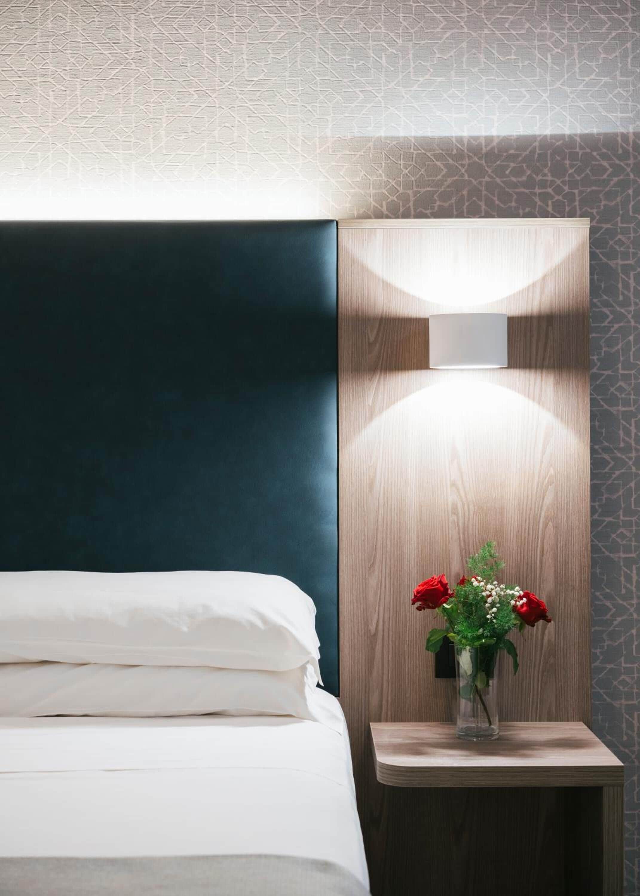 A hotel room with a bed, a nightstand with a vase of red roses, and a wall-mounted light fixture.