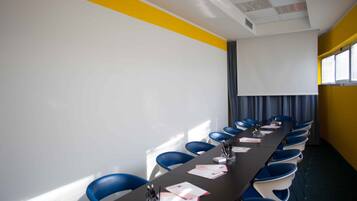 A conference room with a long table, blue chairs, a projector screen, and a yellow accent wall.