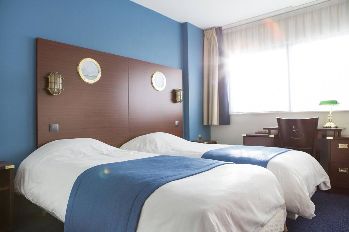 A hotel room with two beds, a wooden headboard, wall-mounted lamps, a desk, and a chair.