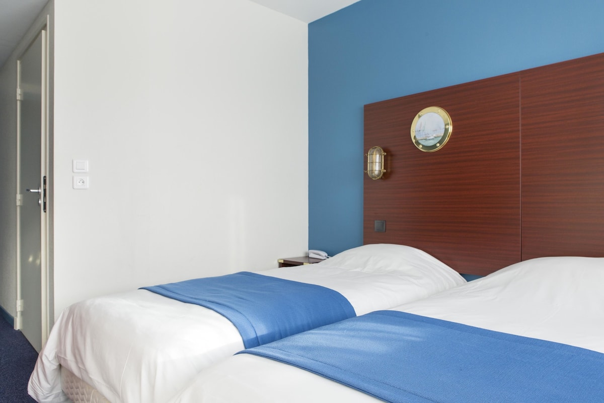 A hotel room with two beds, a wooden headboard, a wall clock, and a bedside table with a lamp.