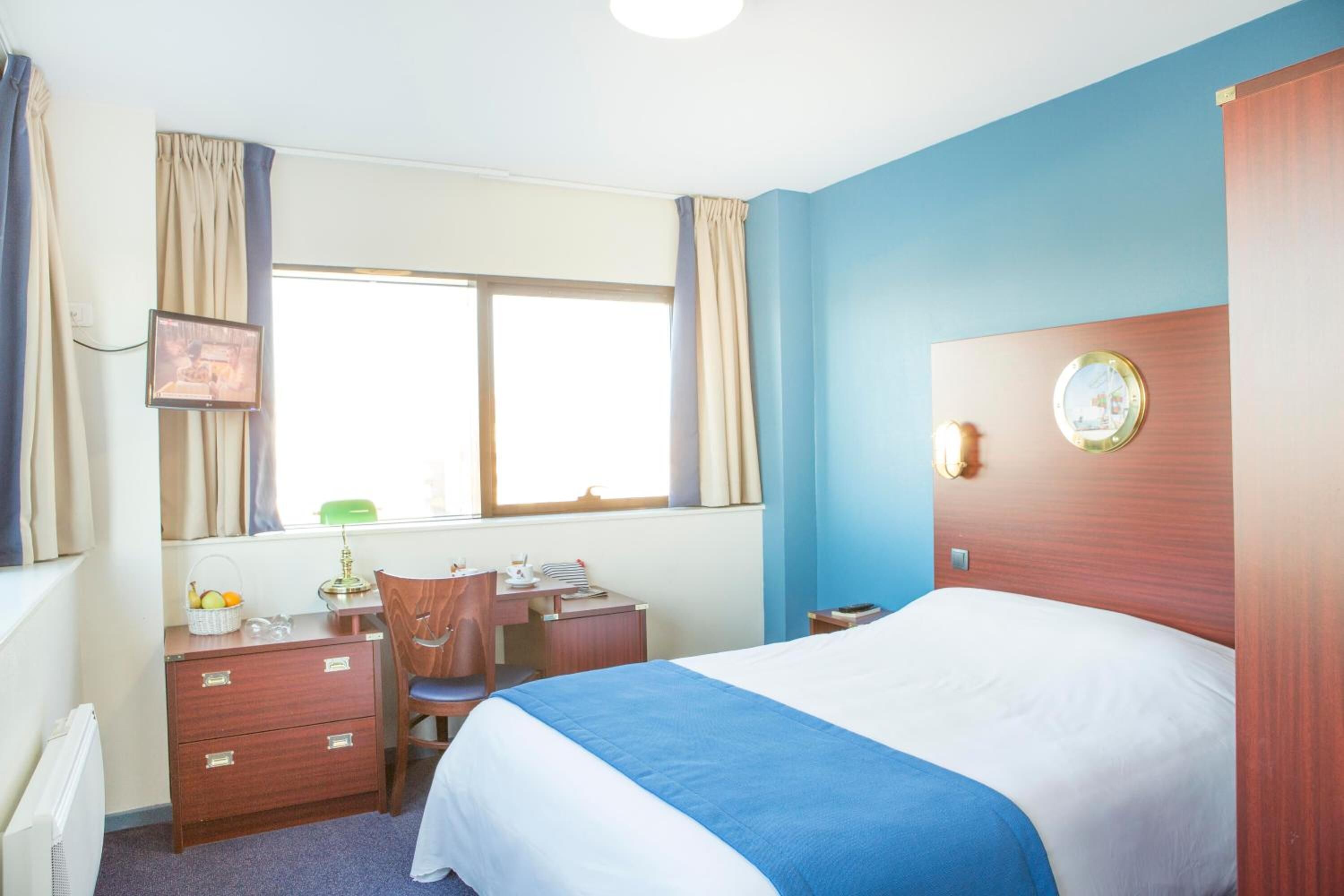 A hotel room with a bed, a desk, a chair, a window with curtains, and a wall clock.