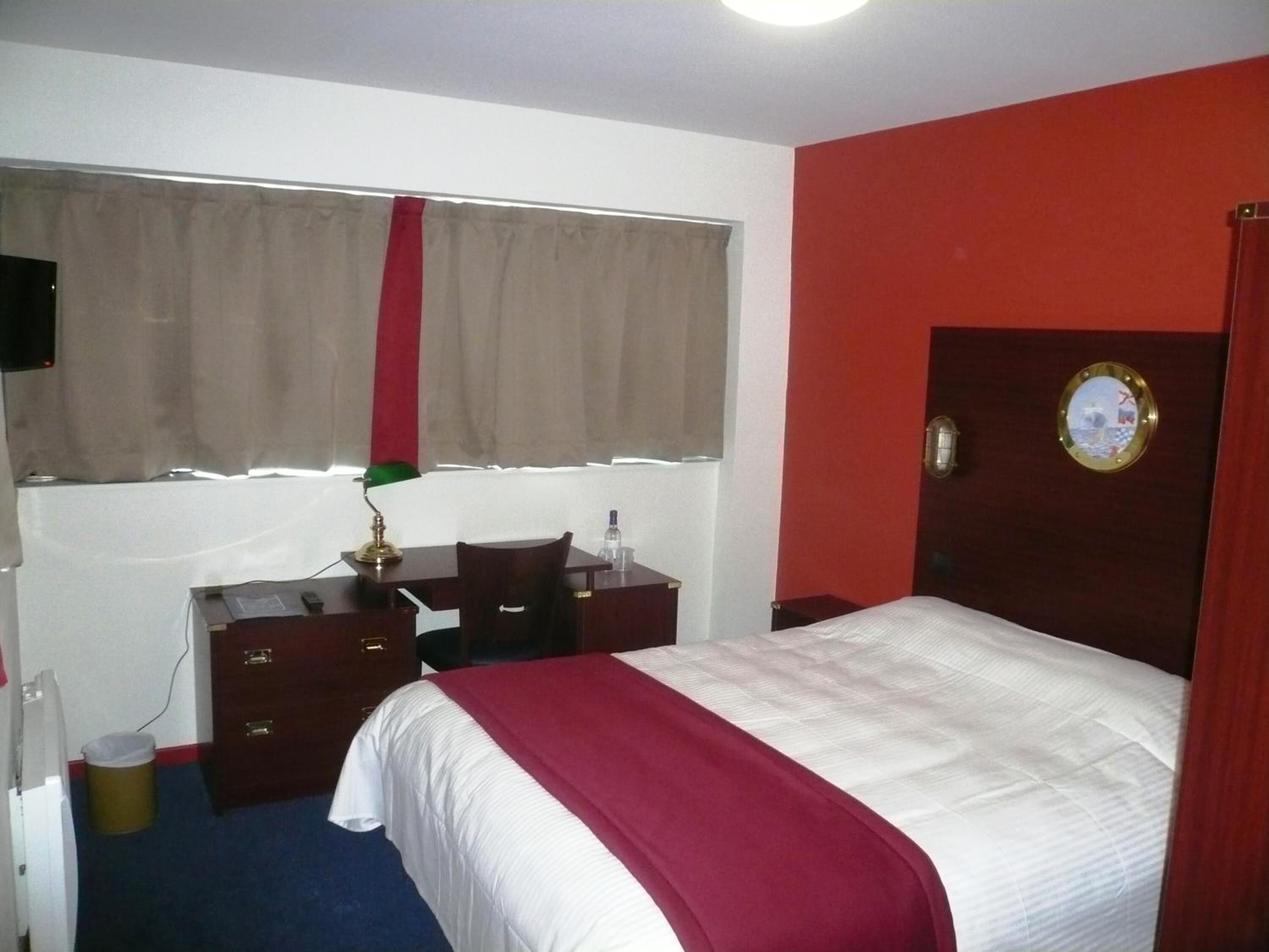 Una habitación de hotel con cama, escritorio, silla, lámpara de escritorio, espejo y ventana con cortinas.