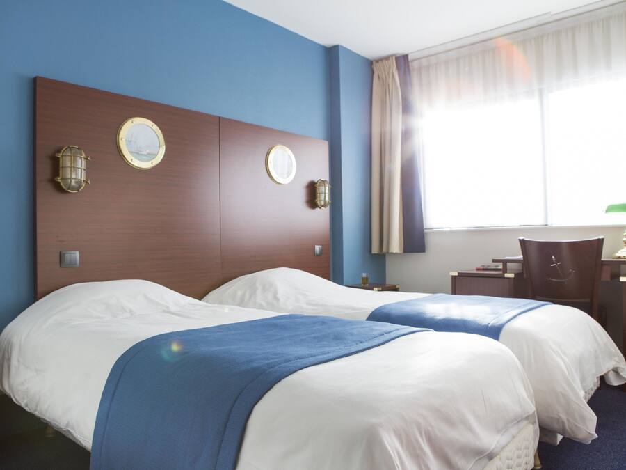 A hotel room with two beds, a wooden headboard, a desk with a chair, and a window with curtains.