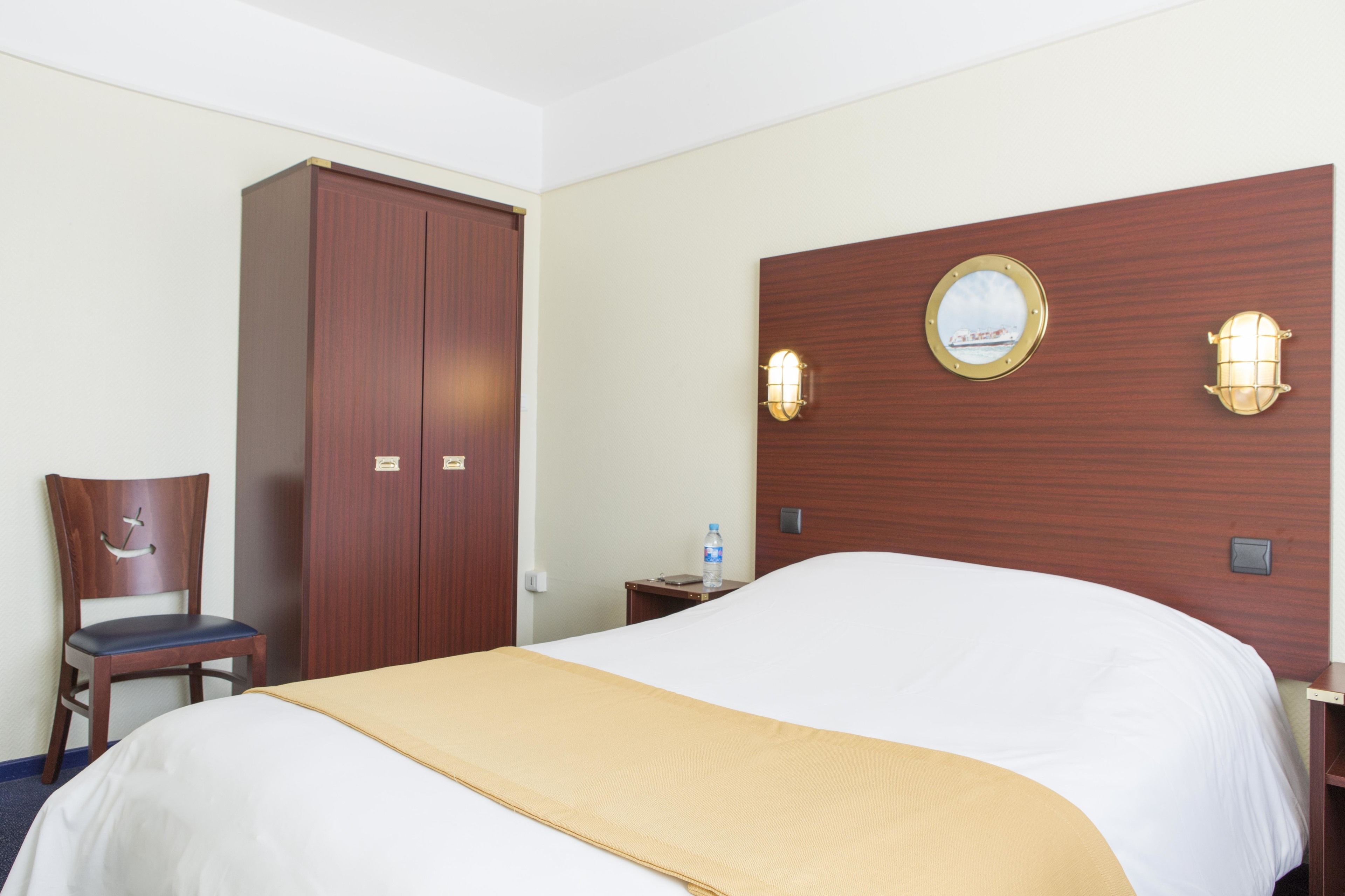 A hotel room with a large bed, a wooden wardrobe, a chair, and a nightstand with a water bottle.