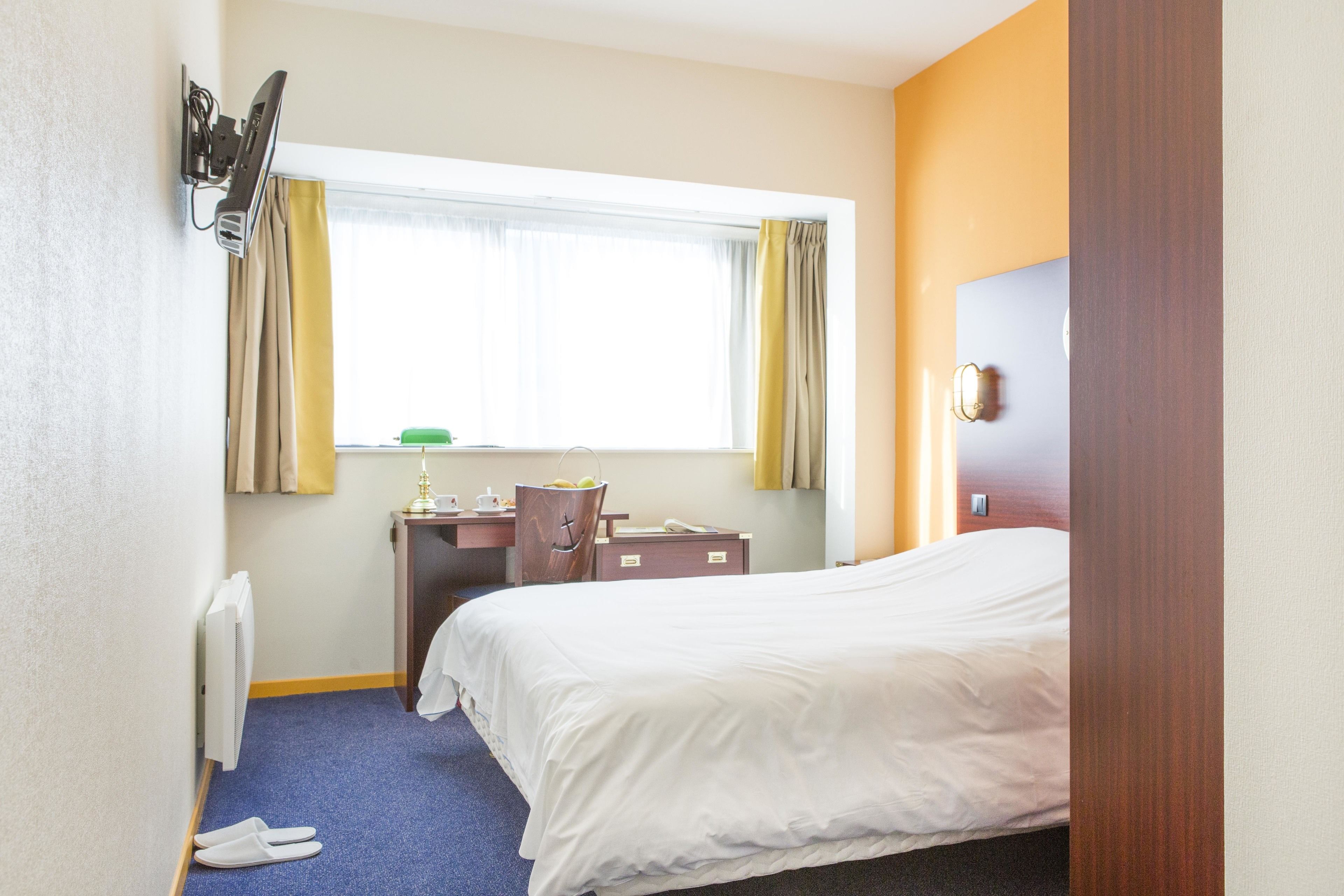 A hotel room with a bed, a desk, a chair, a radiator, and a window with curtains.