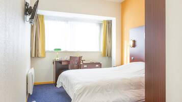 A hotel room with a bed, a desk, a chair, a radiator, and a window with curtains.
