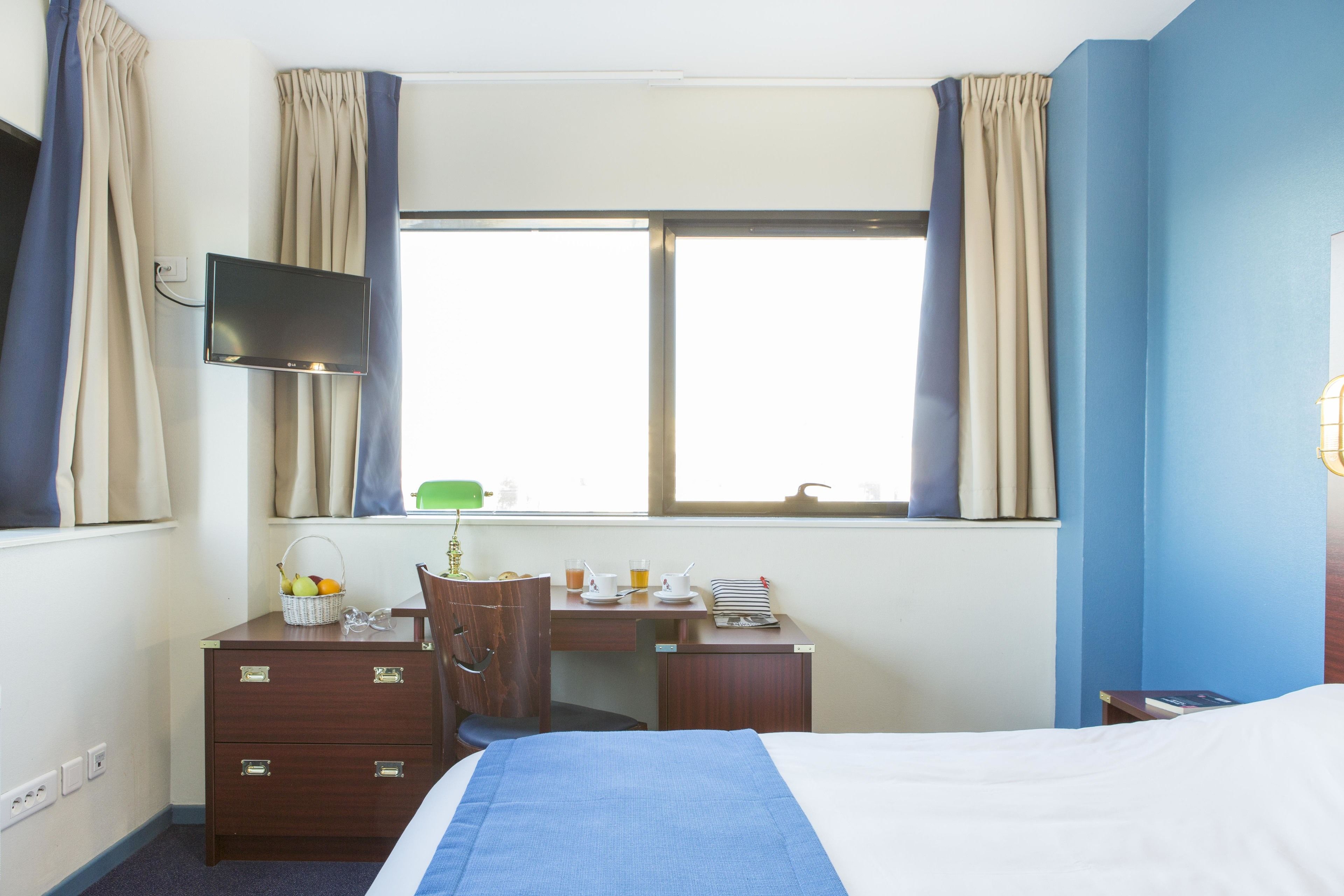 Habitación de hotel con cama, escritorio con computadora, una silla, televisor montado en la pared y ventana con cortinas.