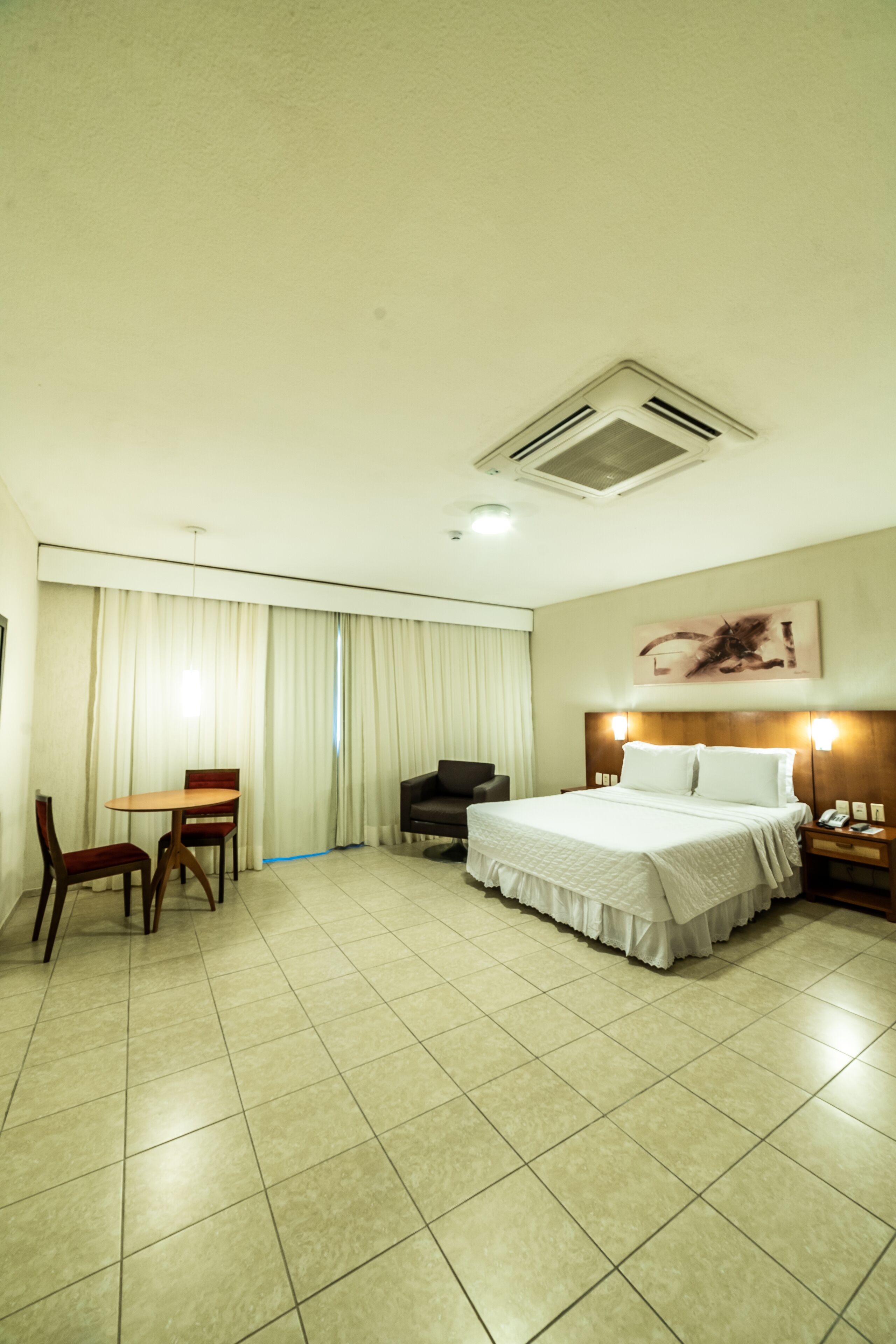 Quarto de hotel com cama, escrivaninha com cadeiras, mesa de cabeceira e um quadro na parede.