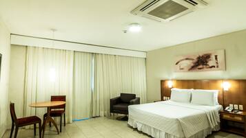 Quarto de hotel com cama, escrivaninha com cadeiras, mesa de cabeceira e um quadro na parede.