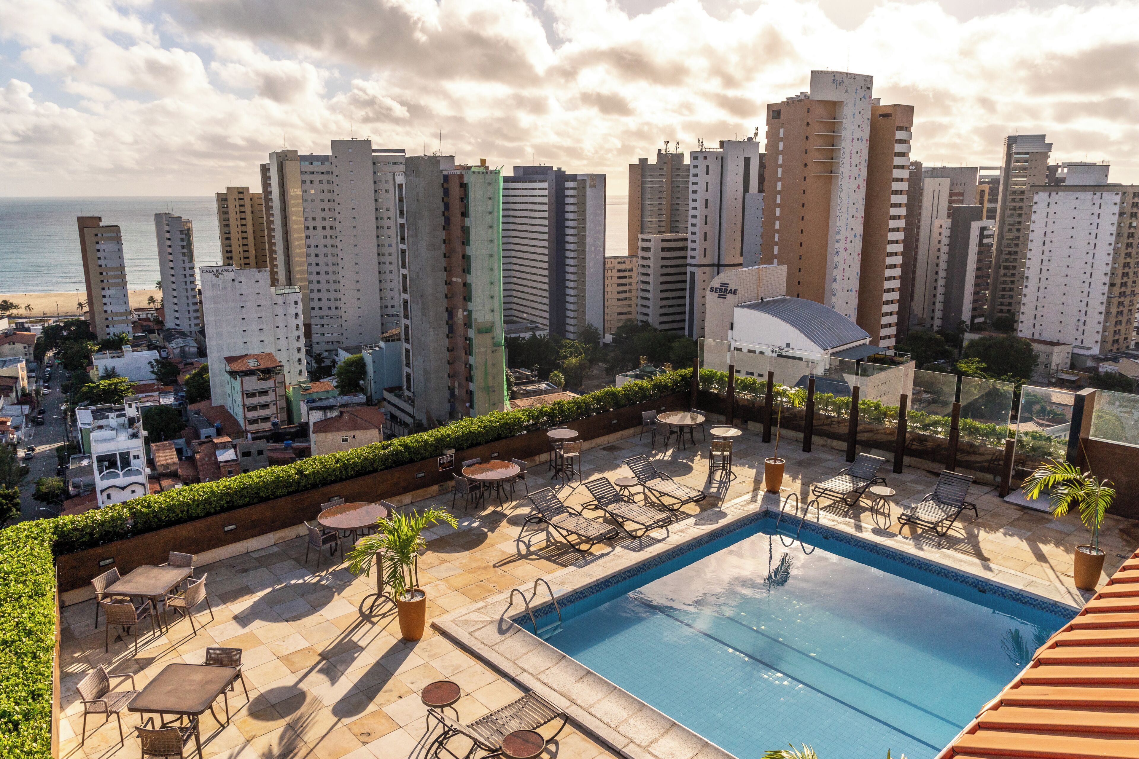A rooftop pool area with lounge chairs, a swimming pool, and a view of tall buildings and the ocean.