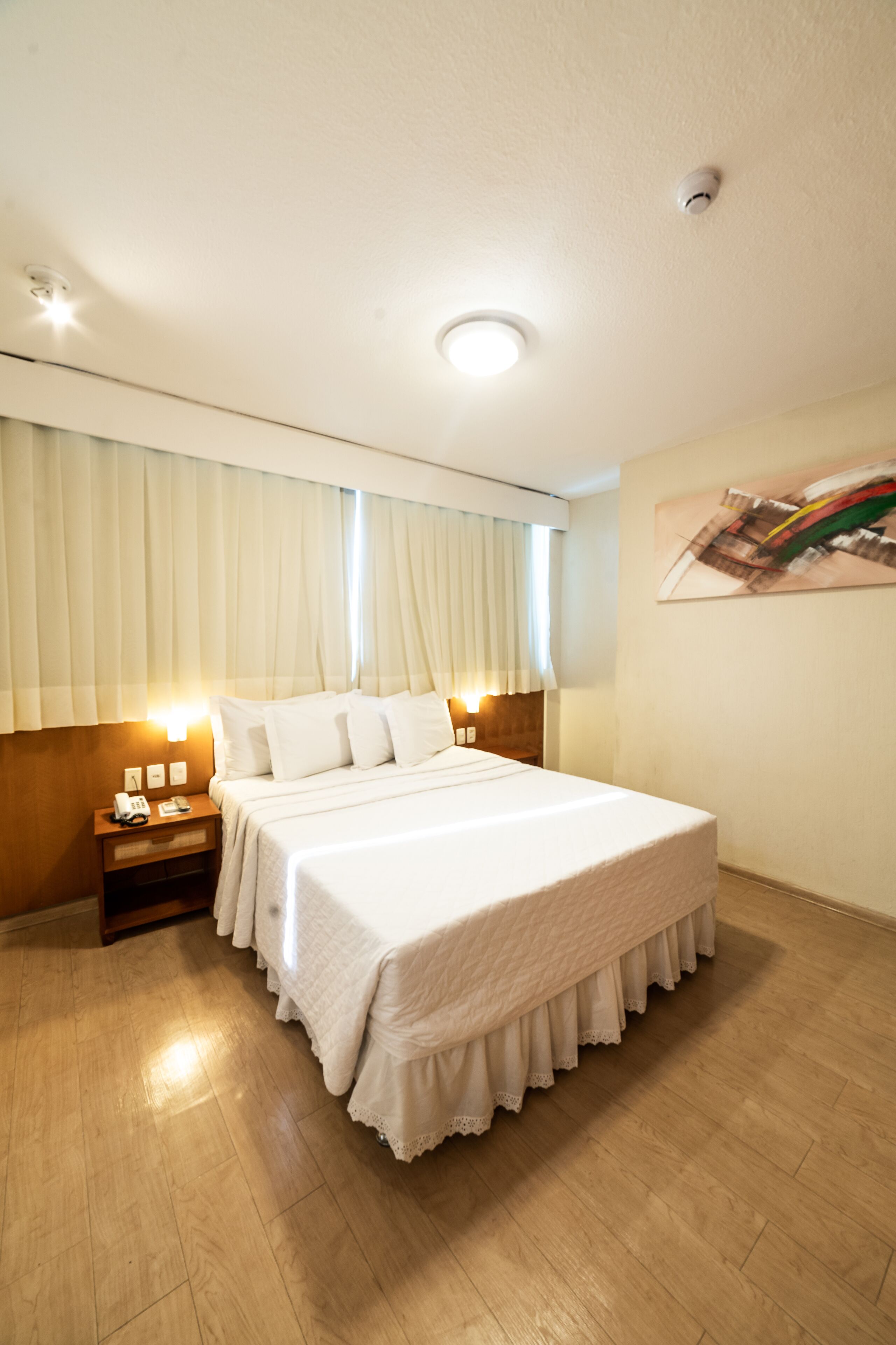 Quarto de hotel bem arrumado, com uma cama grande, mesas de cabeceira e uma luminária no teto.
