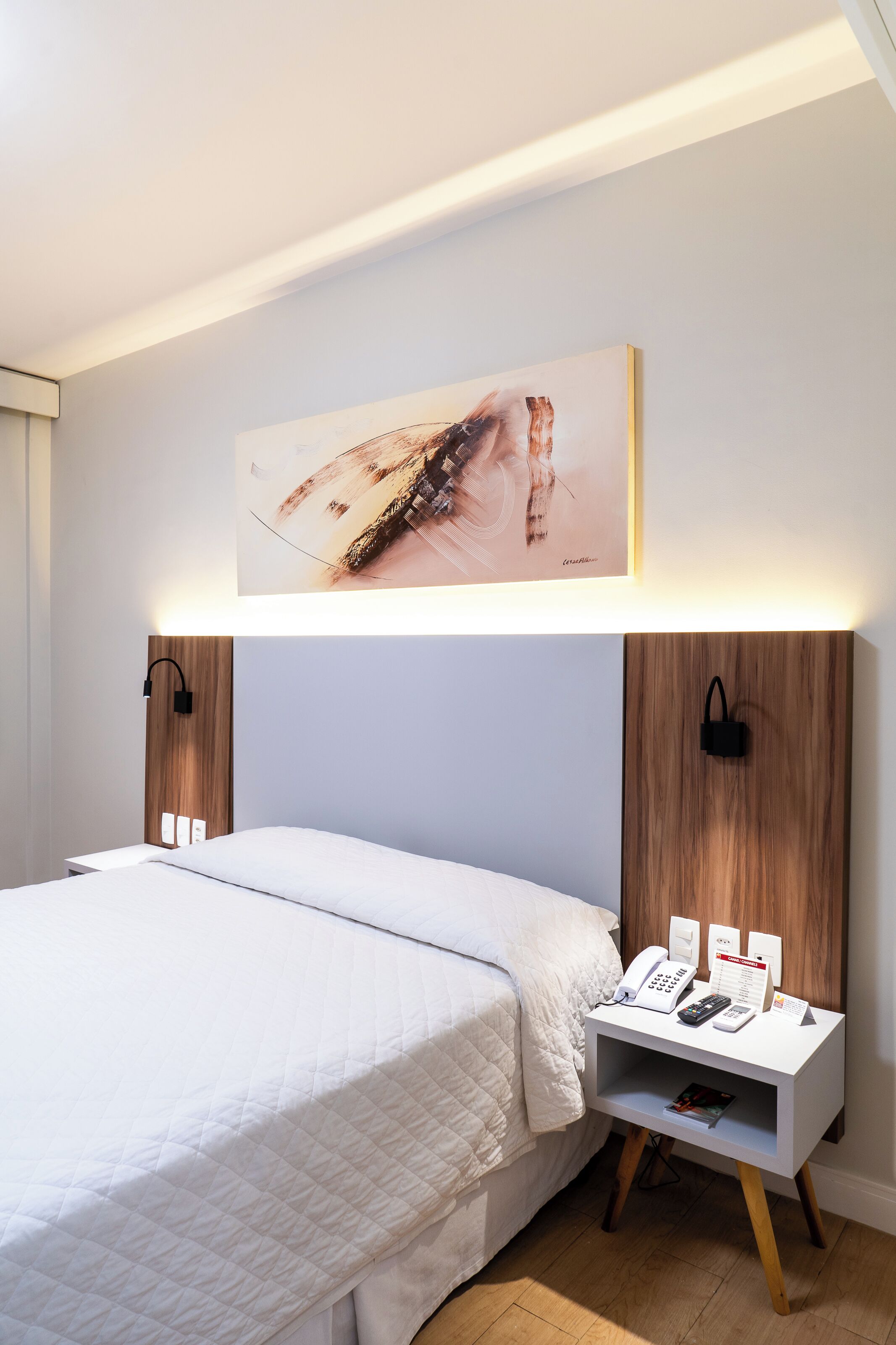 A neatly arranged hotel room with a bed, bedside tables, a wall-mounted artwork, and a wooden headboard.