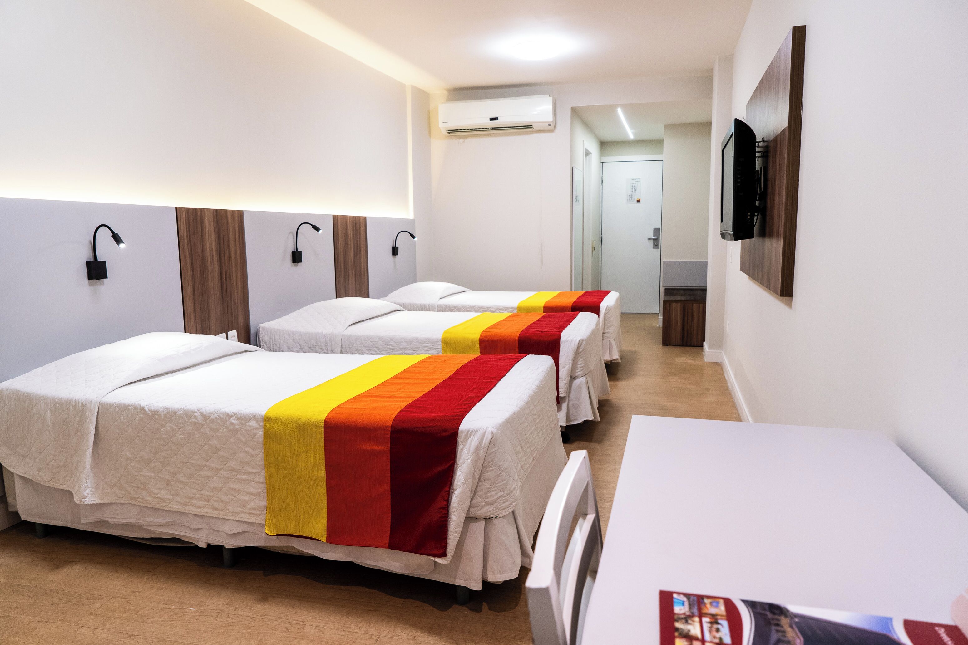 A hotel room with three beds, a TV, an air conditioning unit, and a desk.