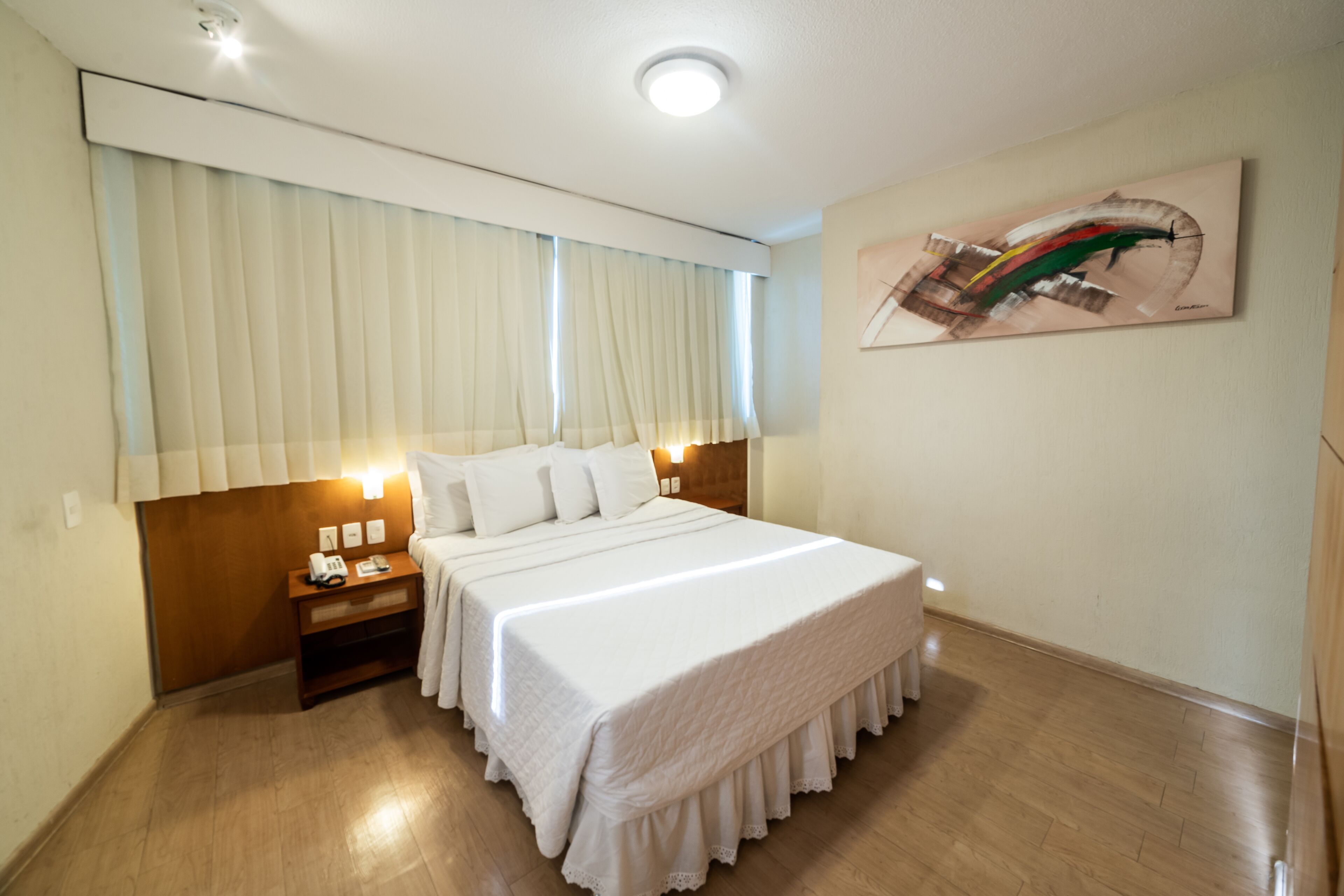 A hotel room with a large bed, wooden headboard, bedside table with a lamp, and a painting on the wall.