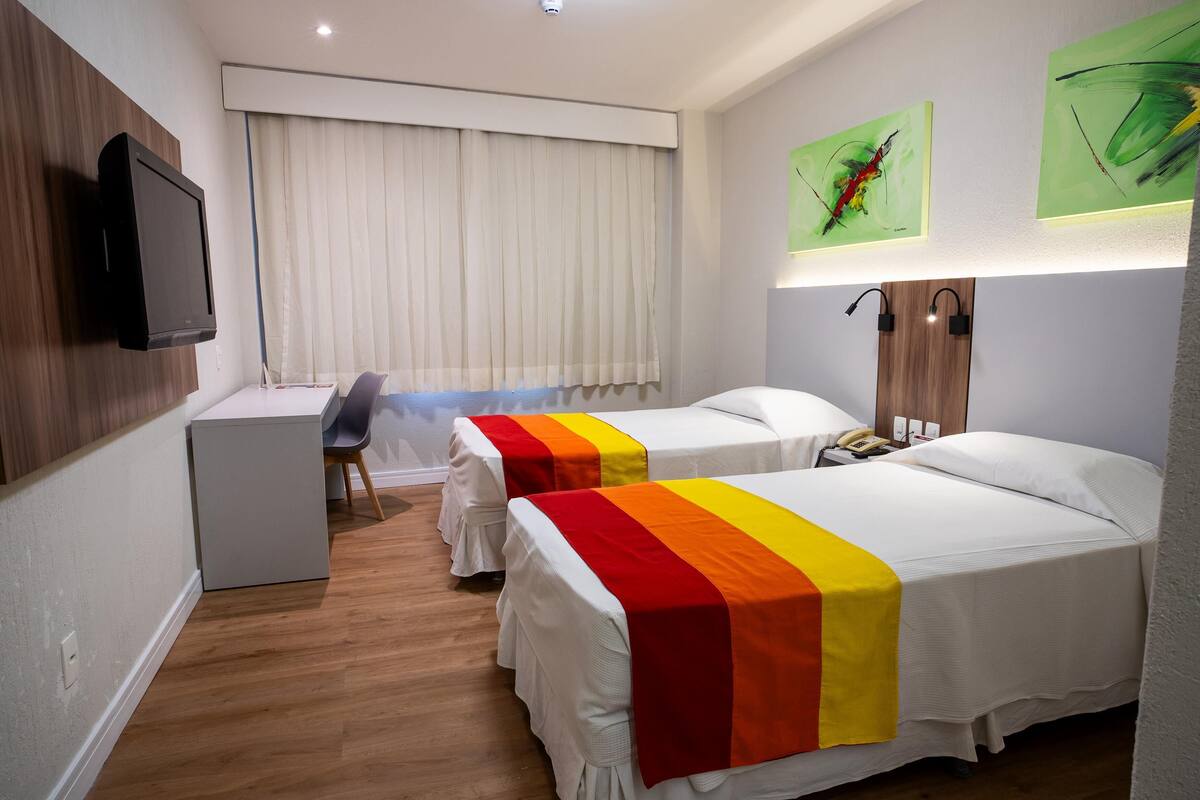 Habitación de hotel con dos camas, un escritorio, una silla, un televisor y obras de arte en las paredes.