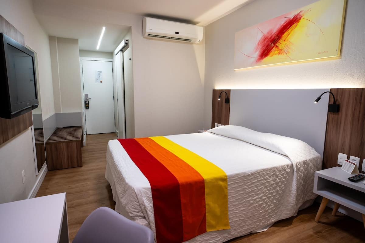 Habitación de hotel con cama, escritorio, televisor, aire acondicionado y un cuadro en la pared.