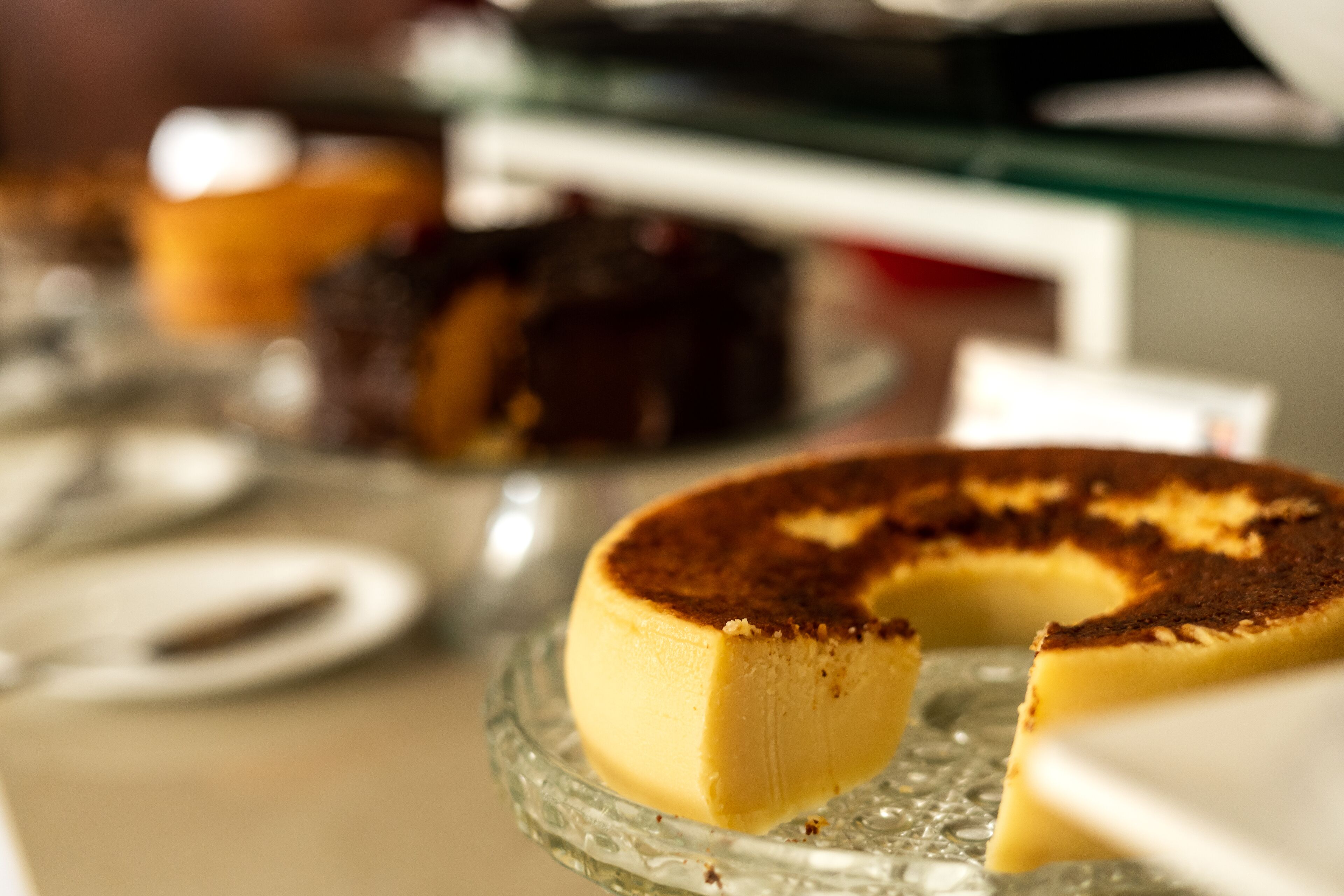 A cheesecake on a glass plate with a slice missing, and a blurred background with other desserts.