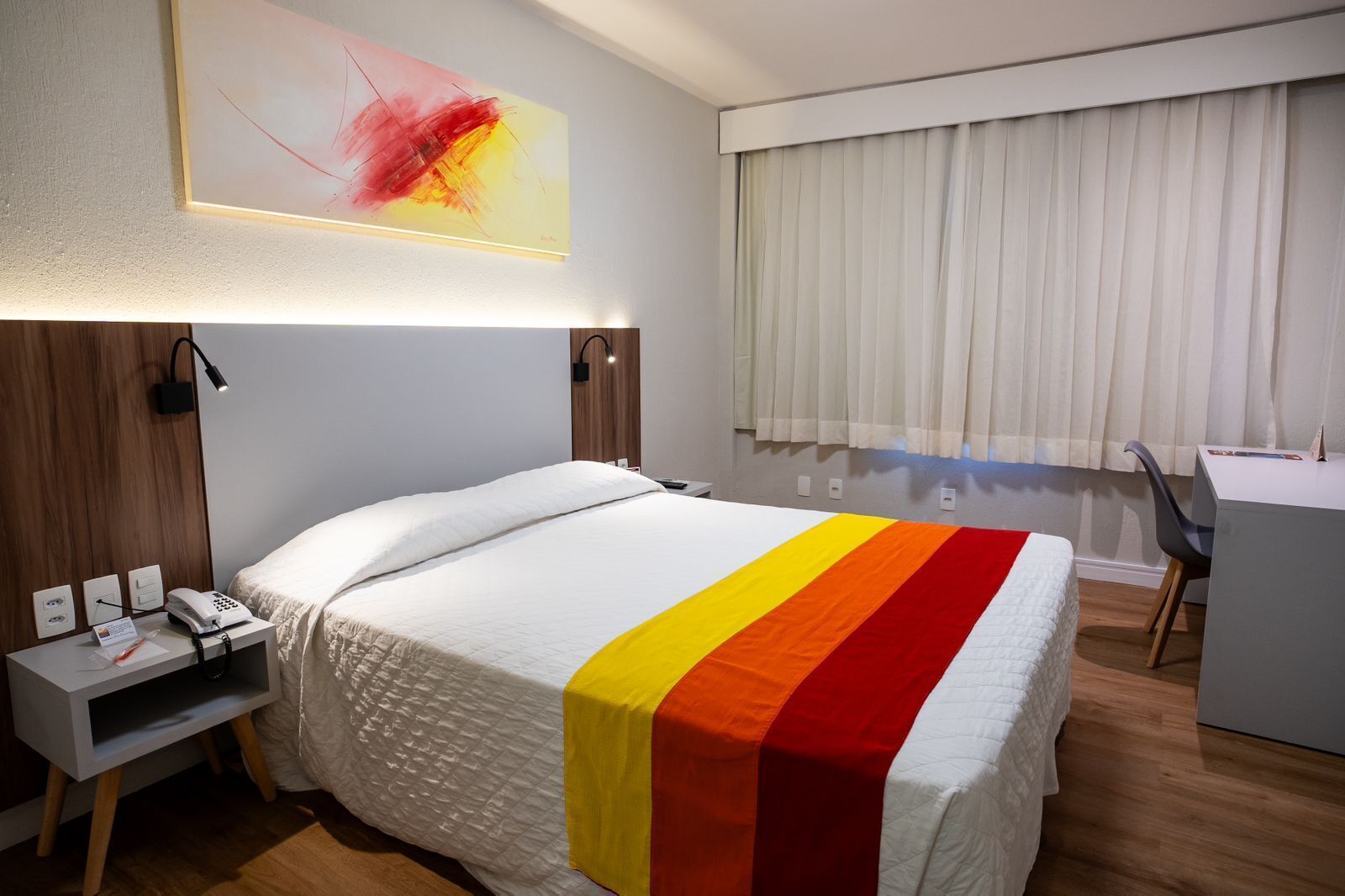 A hotel room with a bed, bedside table, lamp, and a desk with a chair.