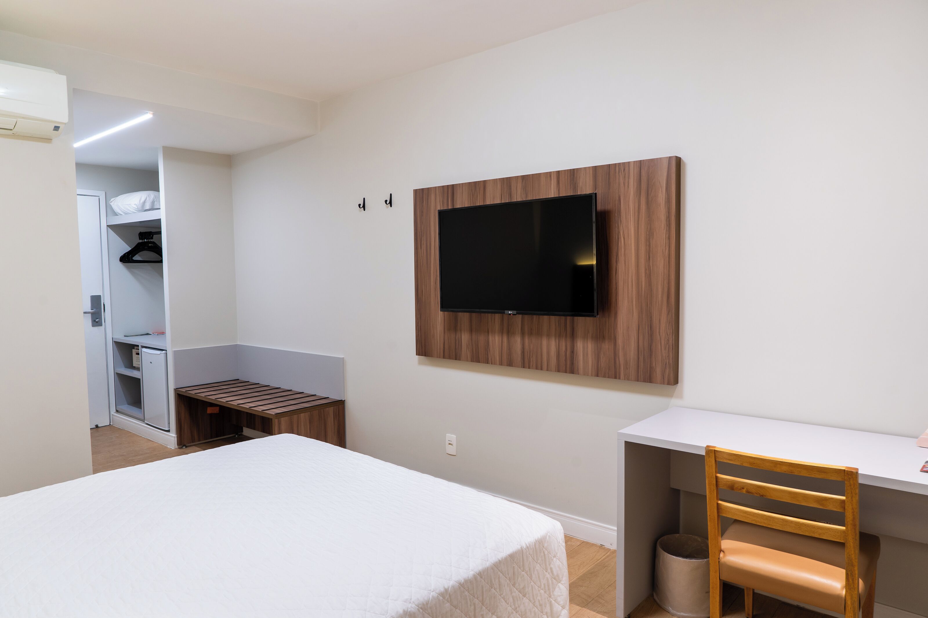 A bedroom with a bed, a desk, a chair, a TV mounted on the wall, and a closet with a hanging rack.