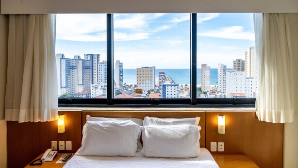 A hotel room with a large window, a bed with white linens, a bedside table with a telephone, and wall-mounted lights. - Hotel Praia Centro (Fortaleza)