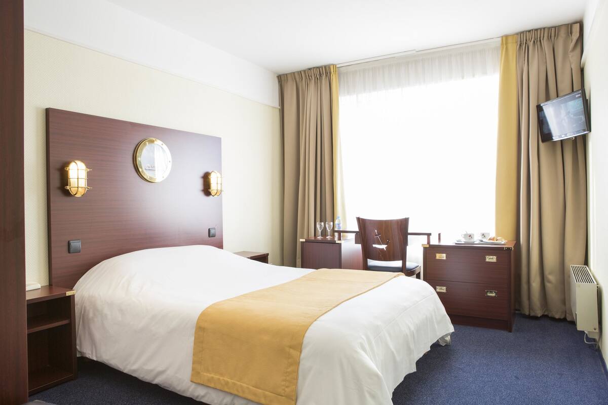 Habitación de hotel con una cama grande, un escritorio con silla, cómoda, televisor de pantalla plana en la pared y ventana con cortinas.