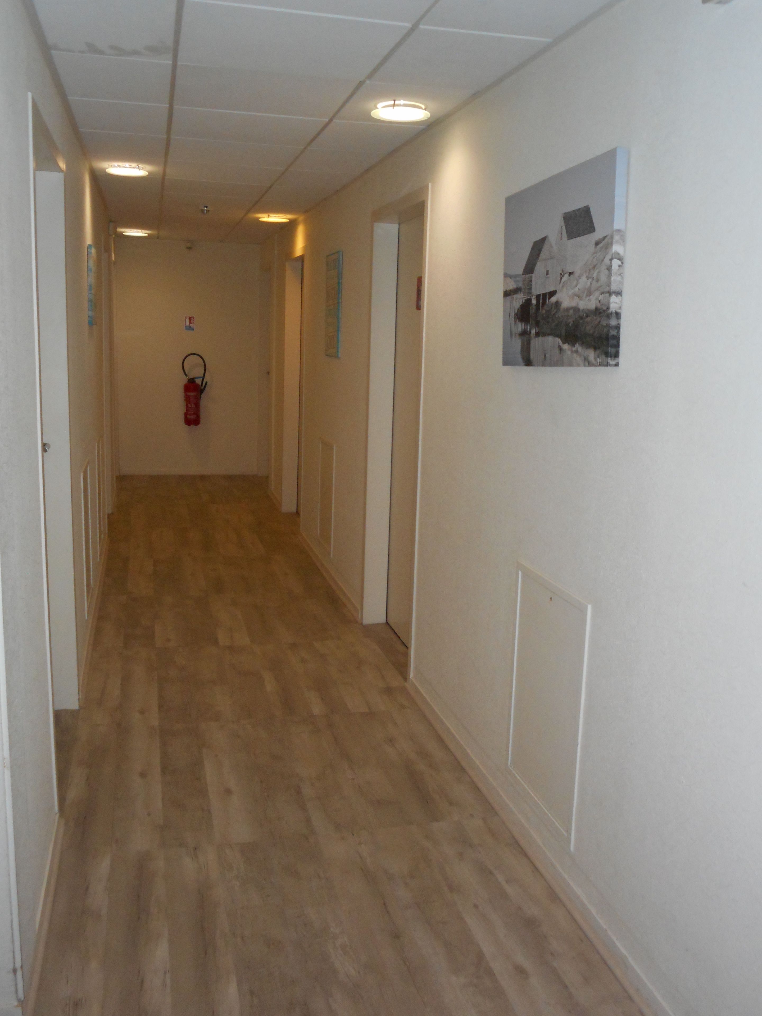 A hallway with wooden flooring, white walls, and doors on both sides. There is a fire extinguisher mounted on the wall and a framed picture on the right wall.