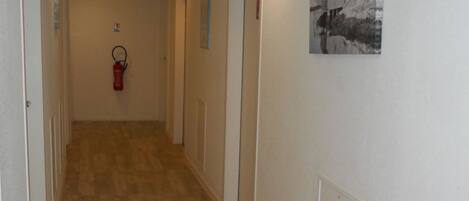 A hallway with wooden flooring, white walls, and doors on both sides. There is a fire extinguisher mounted on the wall and a framed picture on the right wall.