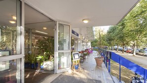 A hotel entrance with glass doors, potted plants, a small easel with a sign, outdoor seating, and a blue barrier.