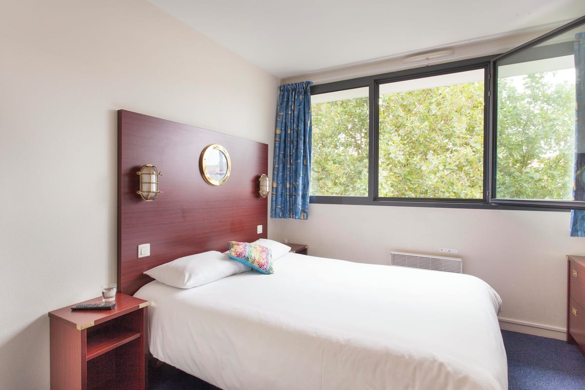 A hotel room with a large bed, a nightstand with a remote control, a wall-mounted lamp, and a window with curtains.