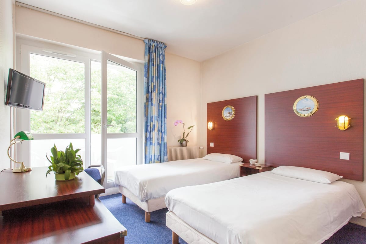 A hotel room with two single beds, a desk with a computer monitor, a potted plant, and a window with curtains.