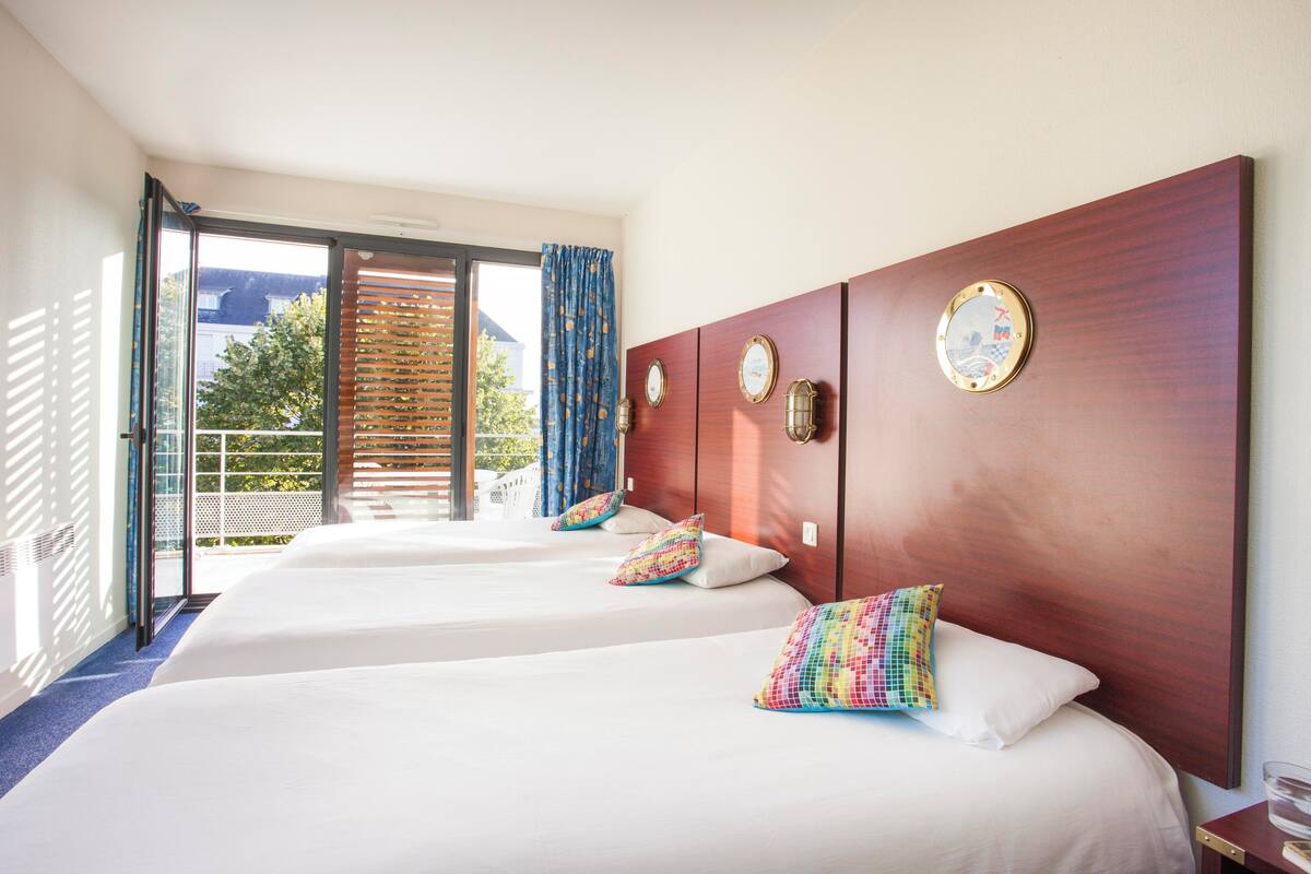 A hotel room with two beds, a wooden headboard, a large window with a view of the outdoors, and a balcony.