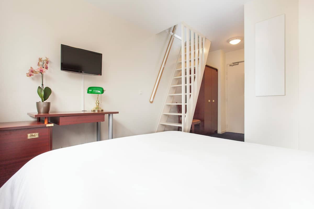 A hotel room with a bed, a desk with a plant, a TV, a green lamp, and a white staircase.