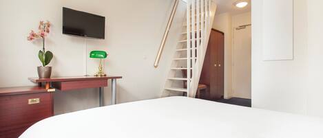A hotel room with a bed, a desk with a plant, a TV, a green lamp, and a white staircase.