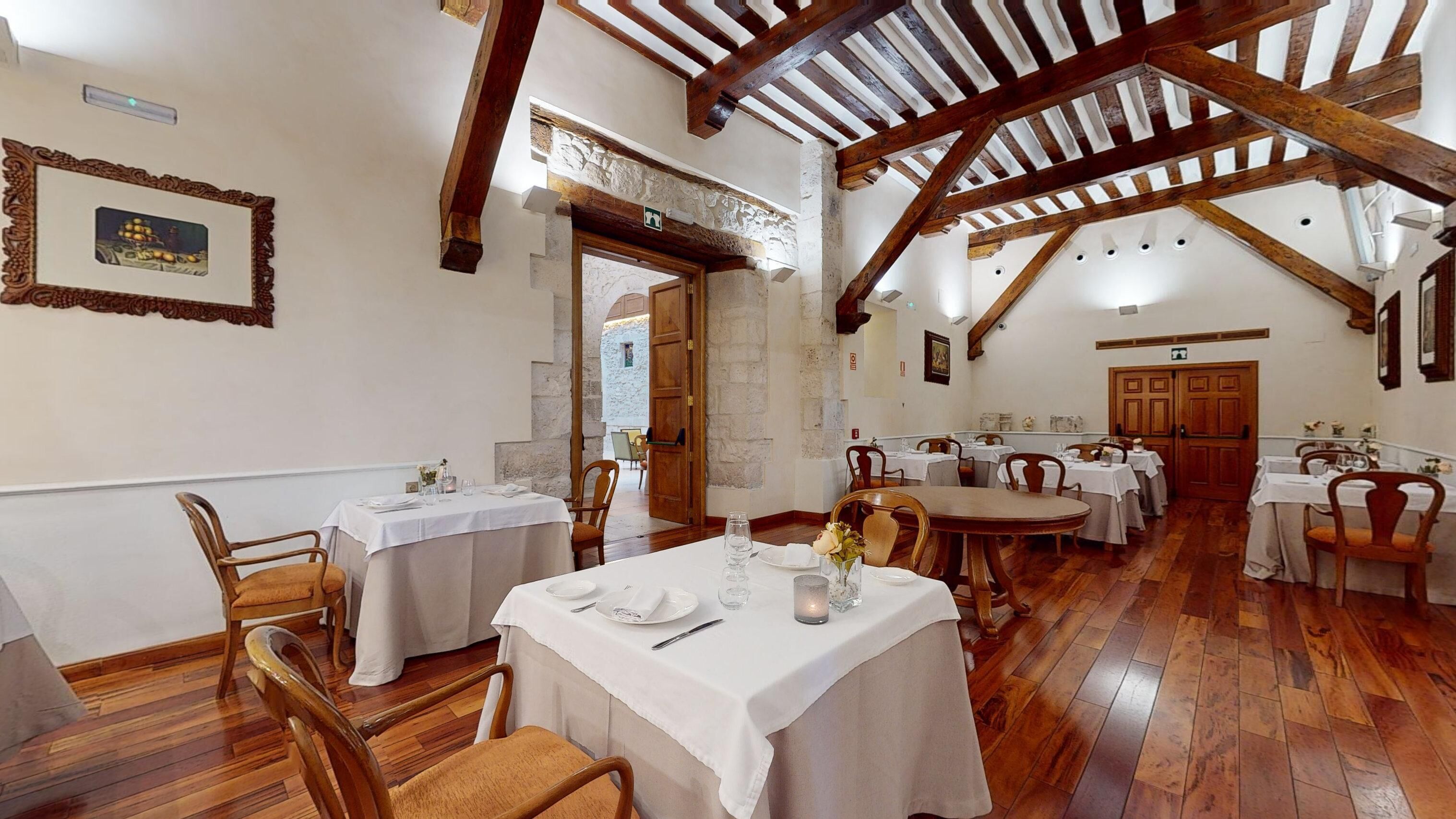 Un restaurant aux poutres apparentes, aux murs blancs et aux éléments décoratifs en pierre. Les tables sont dressées avec des nappes blanches et des chaises.