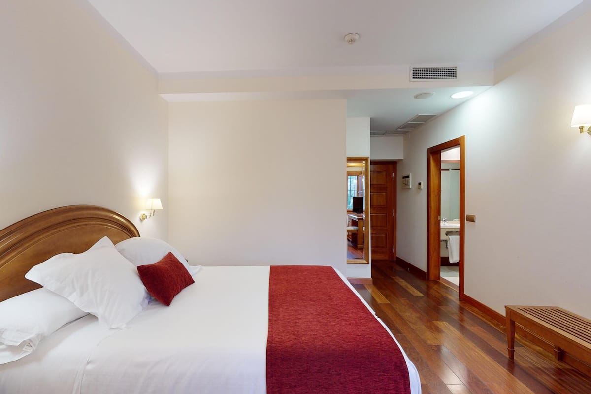 Una habitación de hotel con cabecera de madera, una cama grande con ropa blanca y roja, una manta roja y un banco de madera.