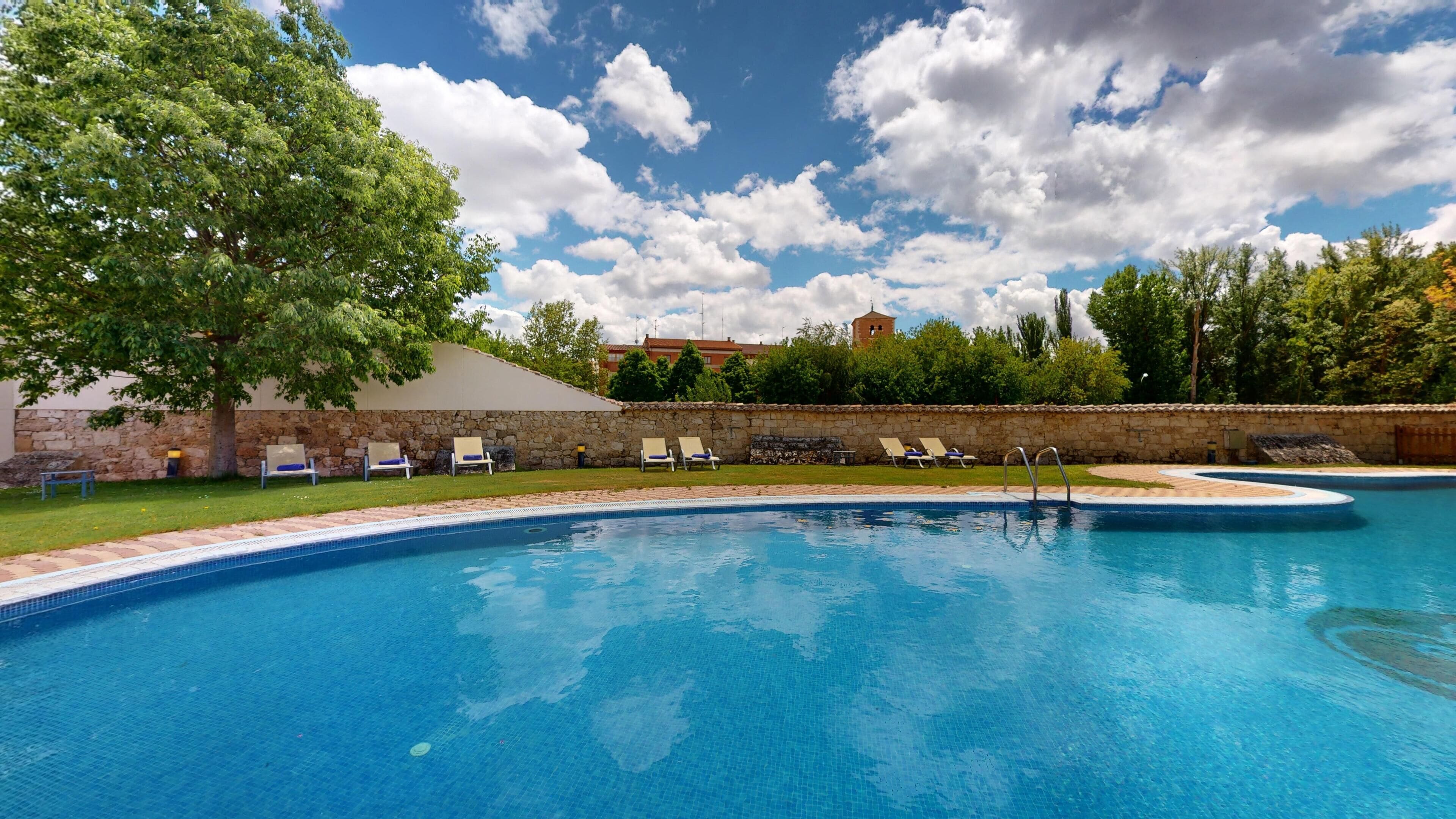 Una piscina con sillones de descanso, un muro de piedra y un edificio al fondo.